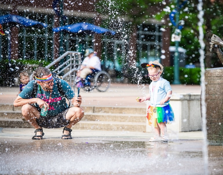 toddler enjoys water fountain while dad watches over her